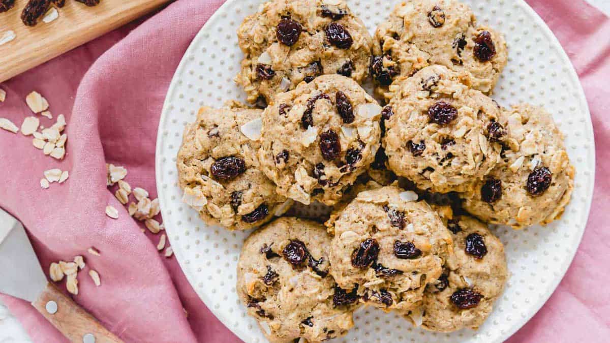 A plate of oatmeal raisin cookies sits on a white dish, placed on a pink cloth, with oats and a spatula nearby.