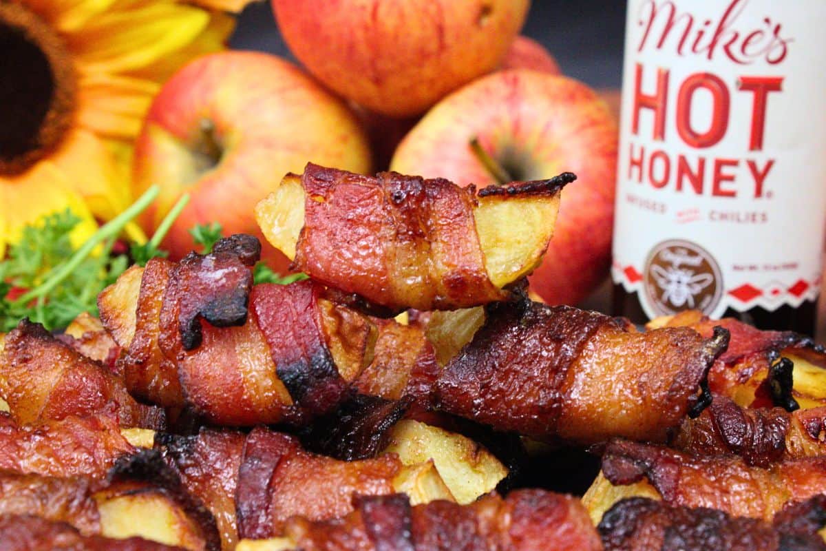 Bacon-wrapped apple slices stacked on a plate, with whole apples, a sunflower, and a bottle of Mikes Hot Honey in the background.