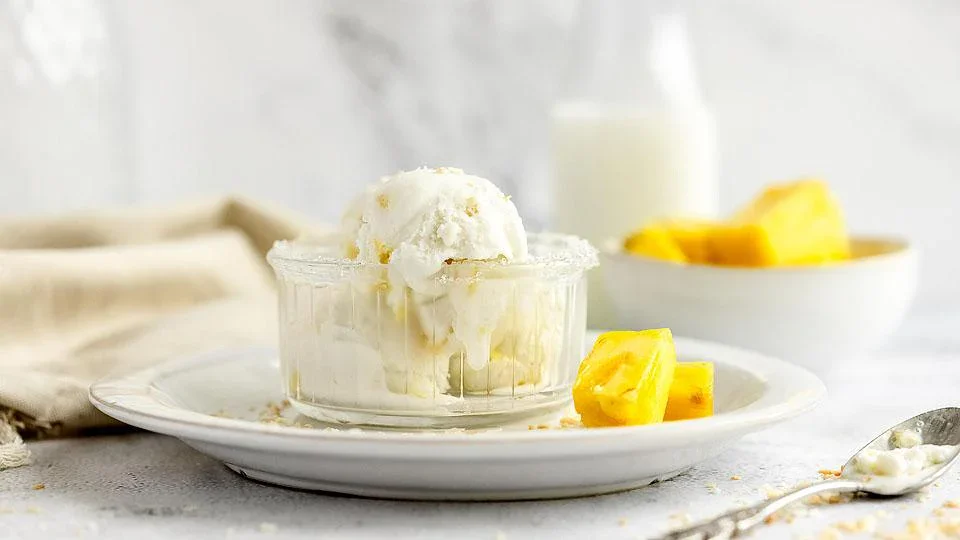 A glass dish of vanilla ice cream sits on a white plate with pineapple chunks, with a bowl of more pineapple and a bottle of milk in the background.