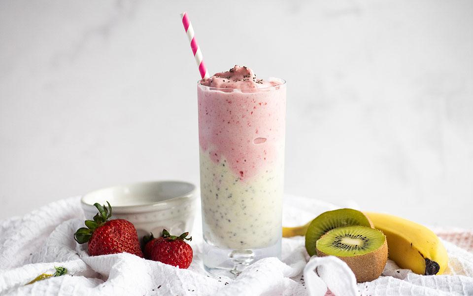 A tall glass with a layered pink and white smoothie, topped with chia seeds and a striped straw. Fresh strawberries, kiwi, and banana are placed beside the glass.