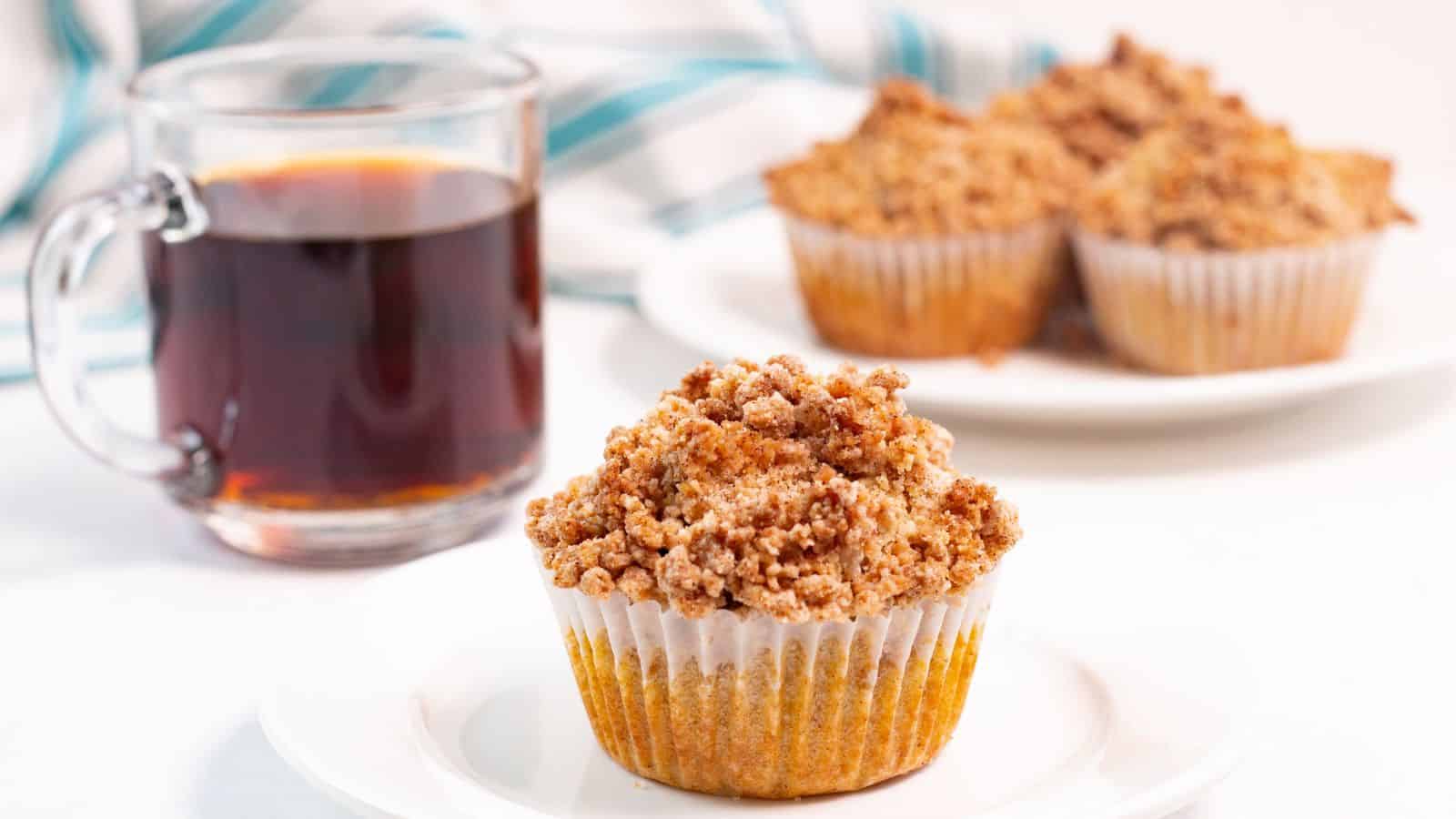 A crumb-topped muffin on a white plate with a glass mug of black coffee. More muffins are on a plate in the background.