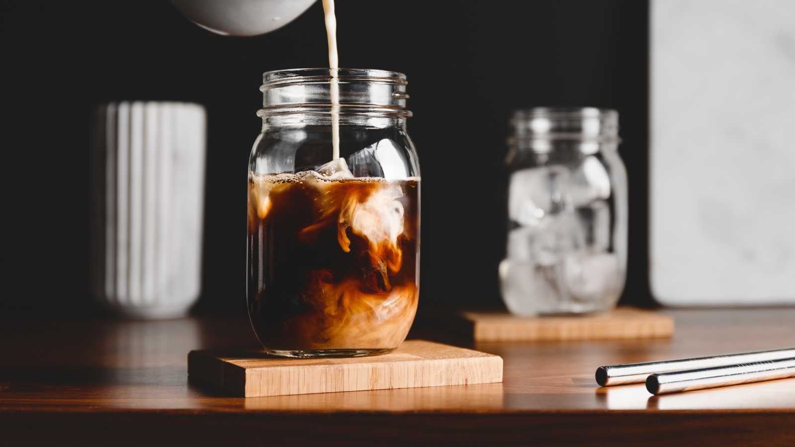 Milk is being poured into a glass jar of iced coffee on a wooden coaster, with another jar of ice and a cup in the background.