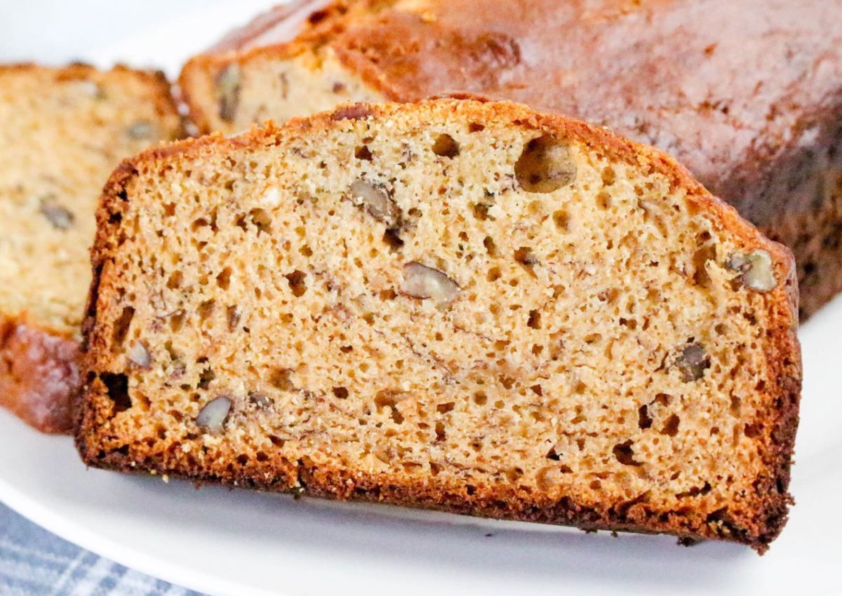 A thick slice of banana bread with visible pieces of walnuts sits on a plate in front of the rest of the loaf.