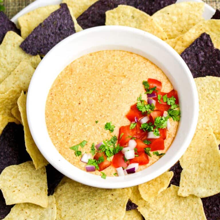 A bowl of creamy cheese dip topped with chopped tomatoes, red onion, and cilantro, surrounded by yellow and blue corn tortilla chips.