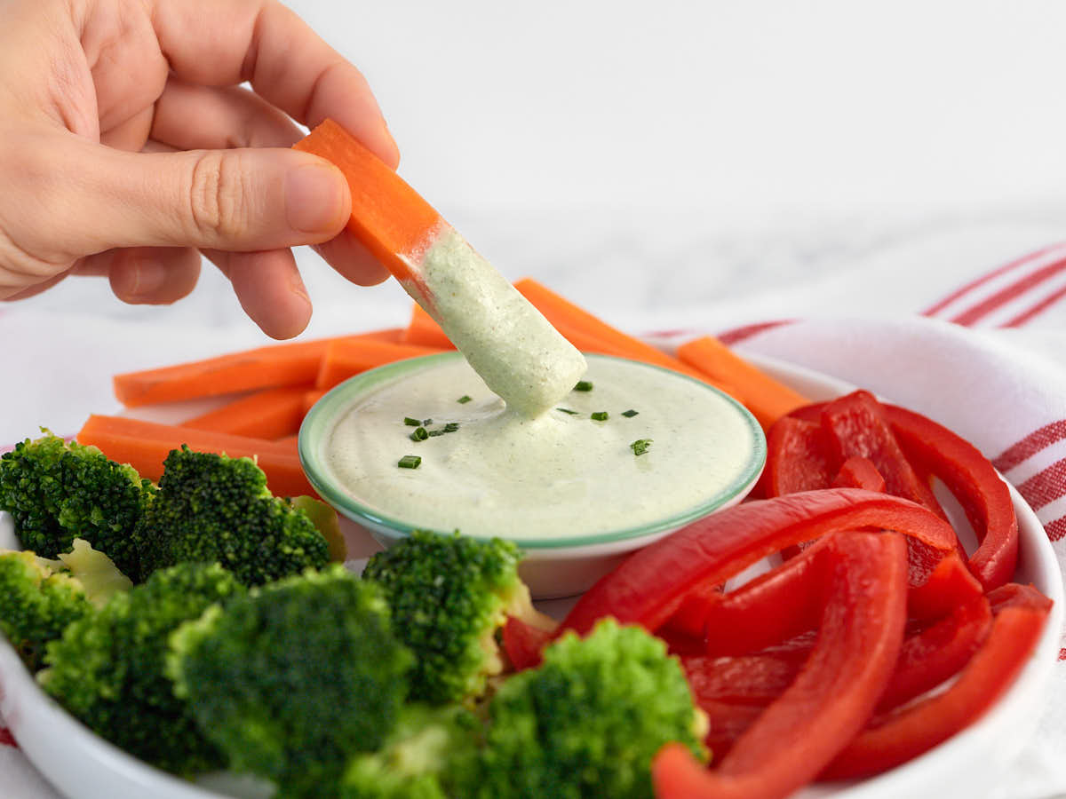 A hand dips a carrot stick into a bowl of creamy dip surrounded by broccoli florets and red bell pepper slices on a white plate.