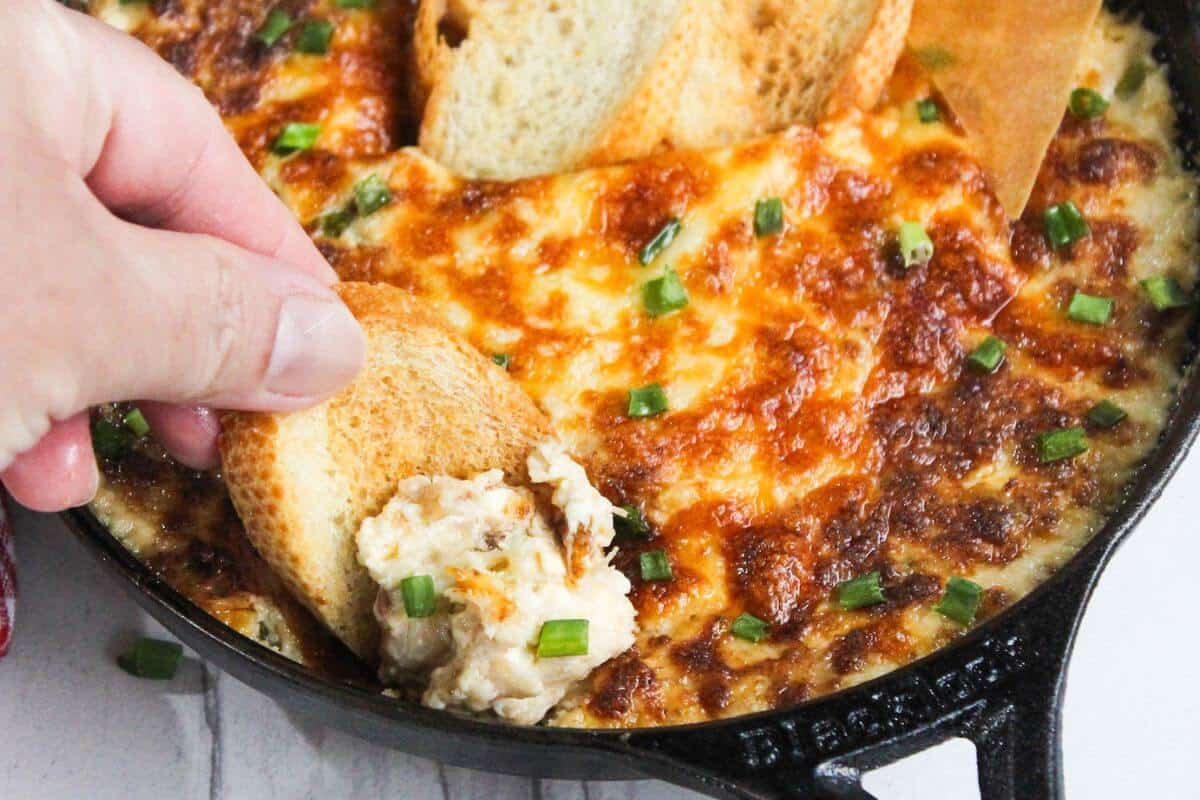 A hand dips a toasted bread slice into a skillet of baked, cheesy dip topped with chopped green onions.