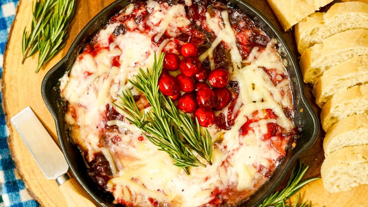 A cast iron skillet filled with melted cheese, cranberries, and herbs, garnished with rosemary, next to sliced bread on a wooden board.