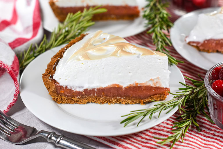 A slice of pie with a graham cracker crust, a red filling, and a thick layer of toasted meringue, served on a white plate with rosemary garnish.