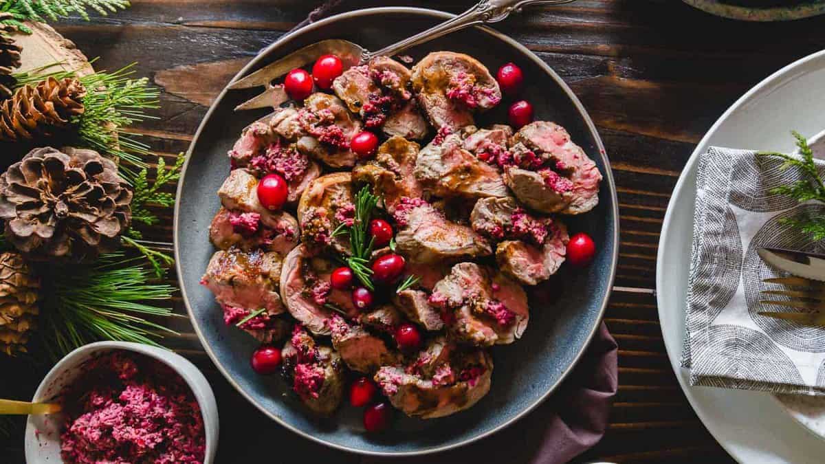A plate of sliced meat garnished with cranberries and herbs is placed on a wooden table with pinecones and a bowl of cranberry sauce nearby.