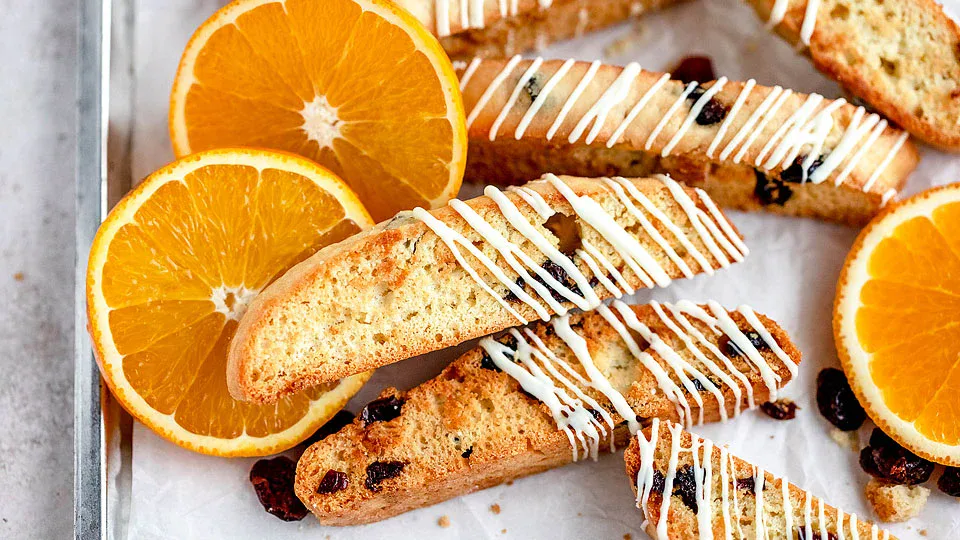 Sliced biscotti with dried fruit and white icing are arranged on parchment paper next to fresh orange slices.