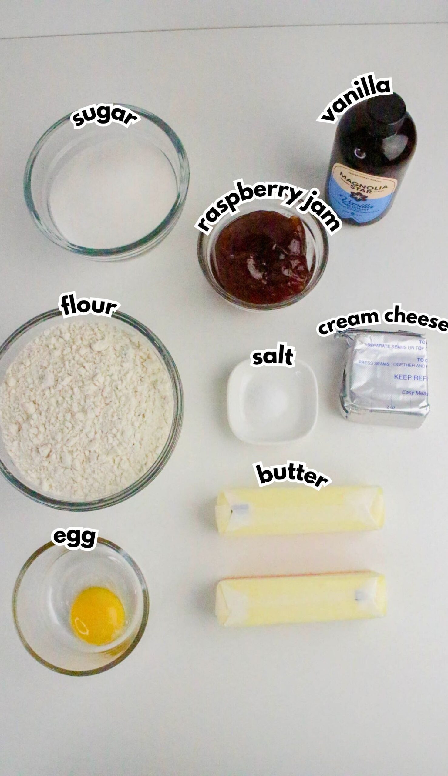 Top-down view of baking ingredients in bowls and packaging: sugar, raspberry jam, vanilla, flour, salt, cream cheese, butter, and an egg on a white background.