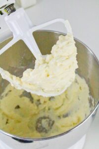 A stand mixer with a paddle attachment holds creamed butter and sugar above a mixing bowl, with more mixture inside the bowl.