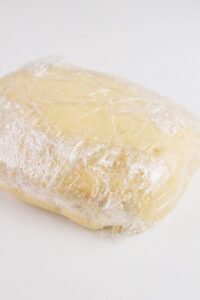 A block of dough wrapped in clear plastic wrap sits on a white surface.
