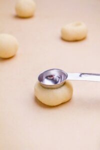 A metal teaspoon presses an indentation into a round ball of dough on a beige surface, with more dough balls in the background.