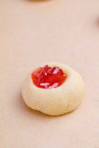 A single round shortbread cookie with a thumbprint center filled with red jam, placed on a light beige surface.