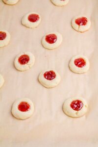 Rows of round shortbread cookies with a dab of red jam in the center are arranged on a baking sheet lined with parchment paper.