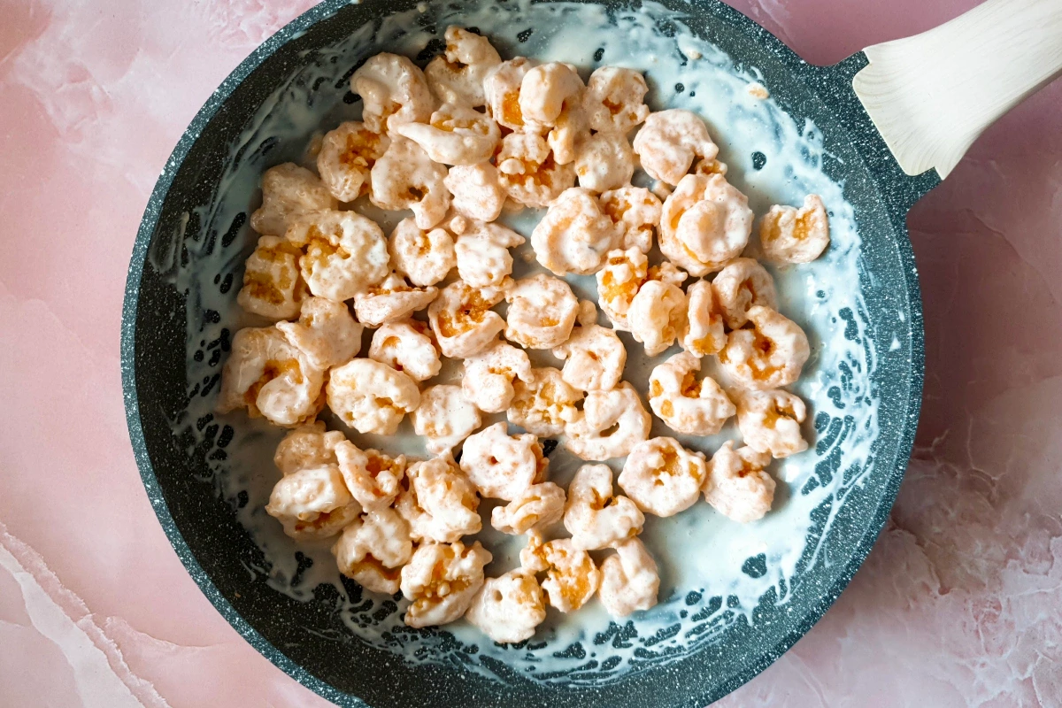 A frying pan filled with shrimp coated in a creamy white sauce, placed on a light pink marble surface.
