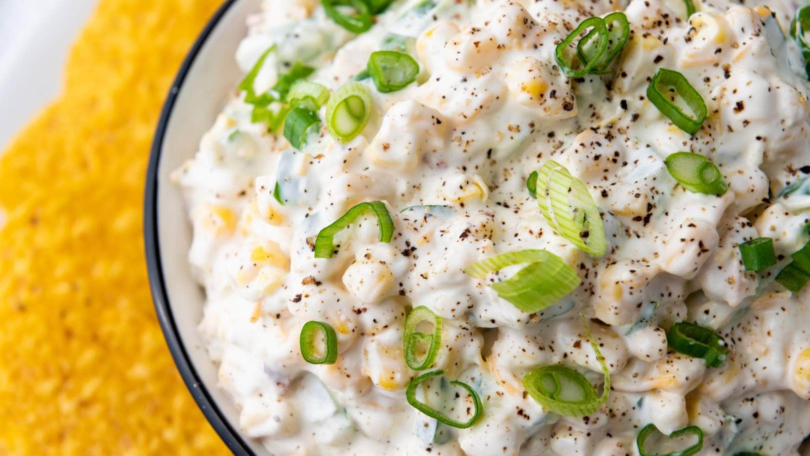 Close-up of a creamy corn salad topped with chopped green onions and cracked black pepper, served in a white bowl with tortilla chips on the side.