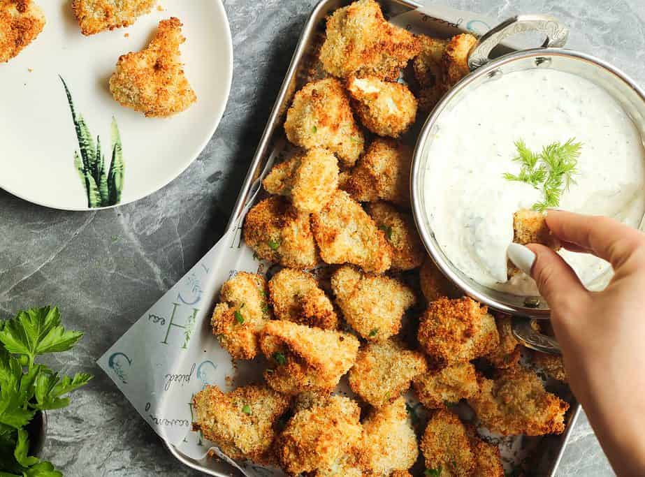 A tray of breaded and baked cauliflower bites is served with a bowl of creamy dip; a hand dips one piece into the sauce. A plate with cauliflower bites is nearby.