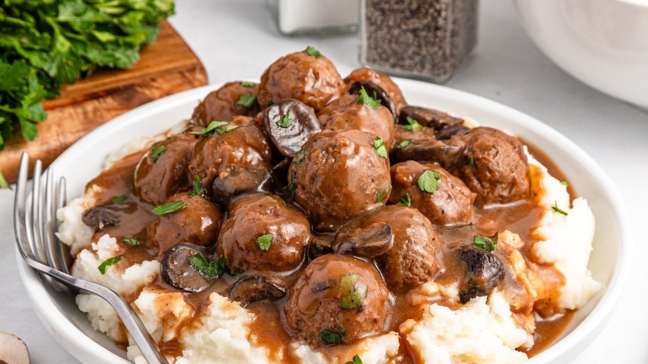 A plate of mashed potatoes topped with meatballs, brown gravy, mushroom slices, and chopped parsley, with a fork on the side.