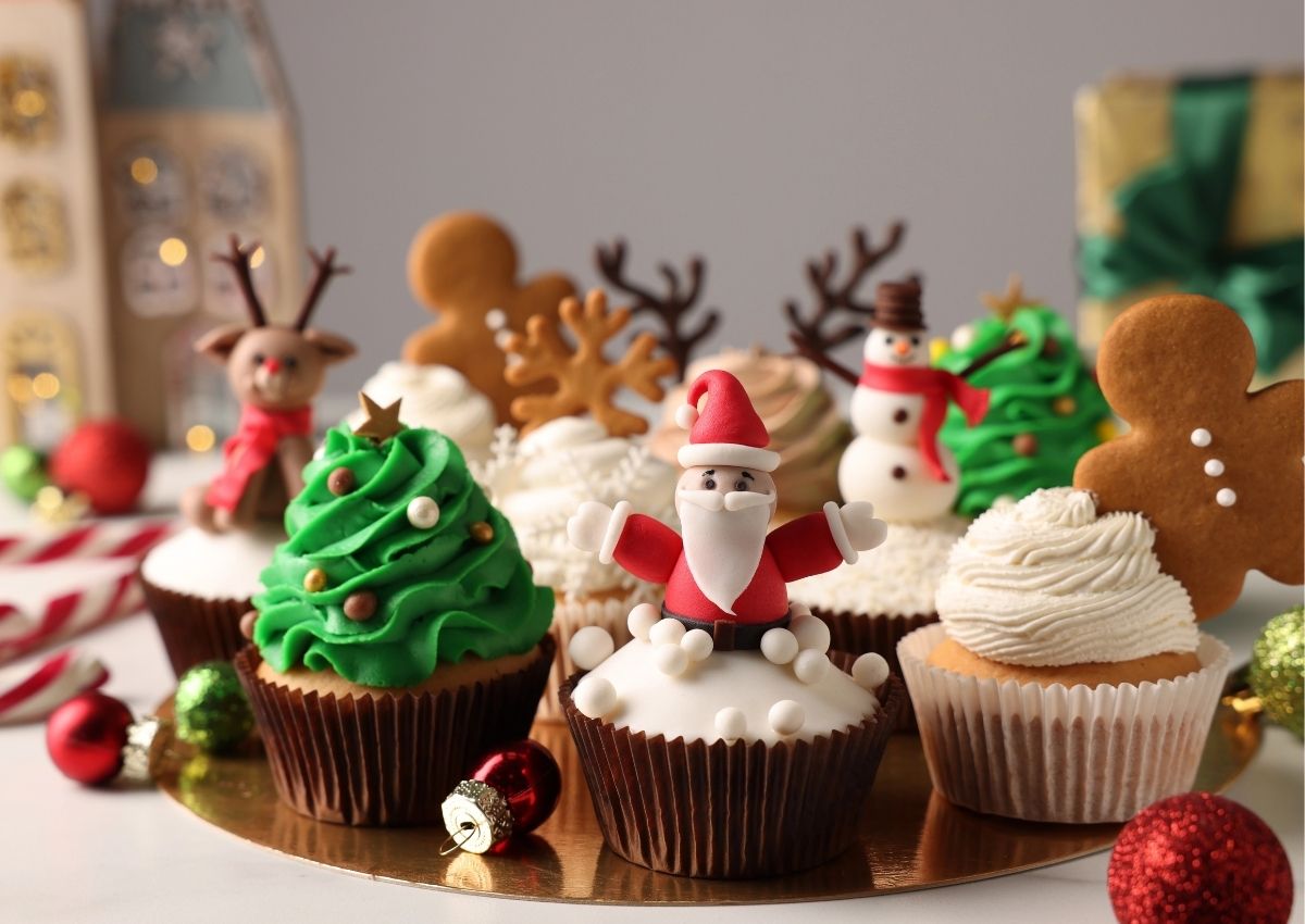 A variety of Christmas-themed cupcakes decorated with fondant figures of Santa, reindeer, snowmen, Christmas trees, and gingerbread men, displayed on a gold platter.