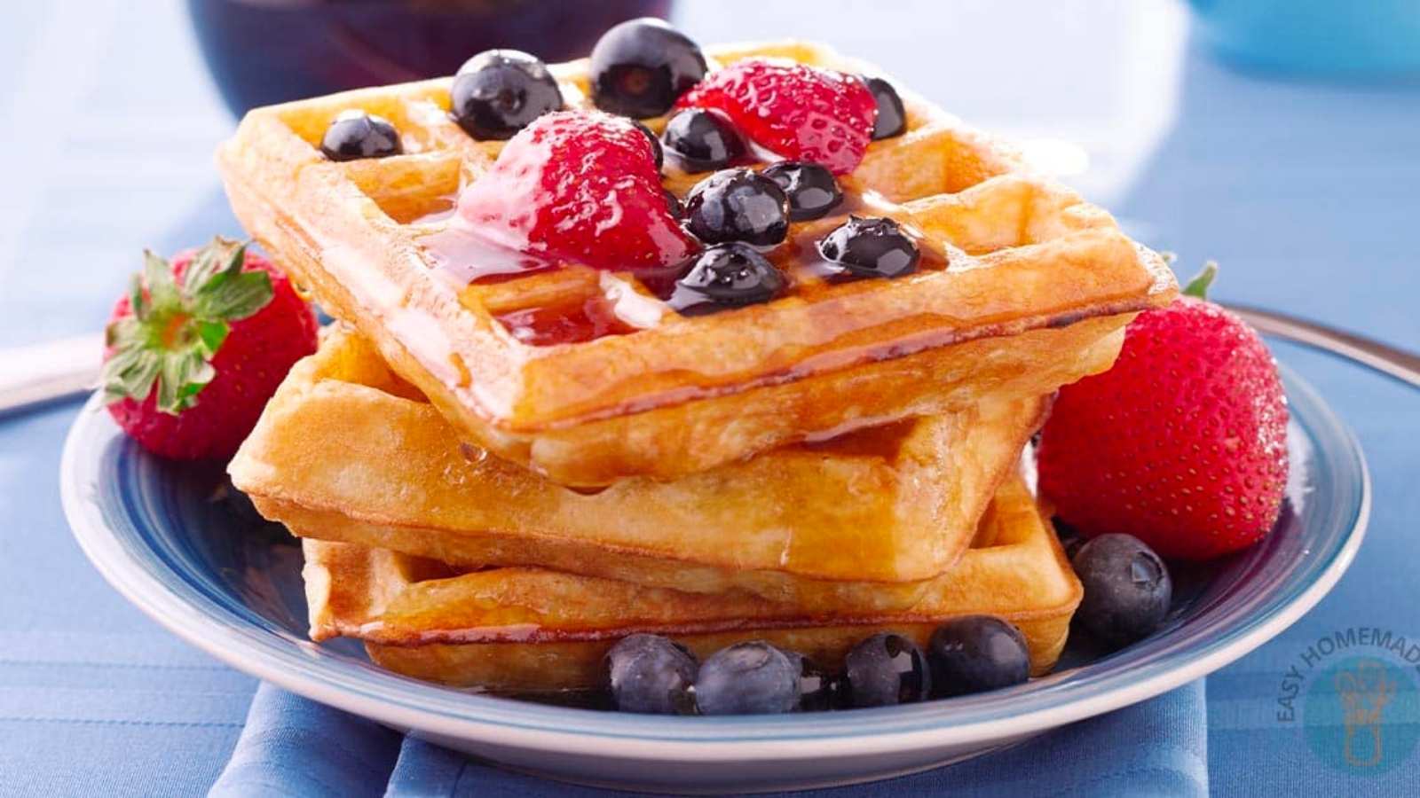 A stack of waffles topped with strawberries, blueberries, and syrup sits on a blue plate with extra berries on the side.