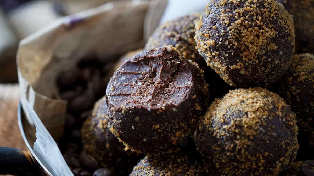 A close-up of chocolate truffles coated with crumbled topping, with one truffle showing a bite taken out to reveal the dense interior.