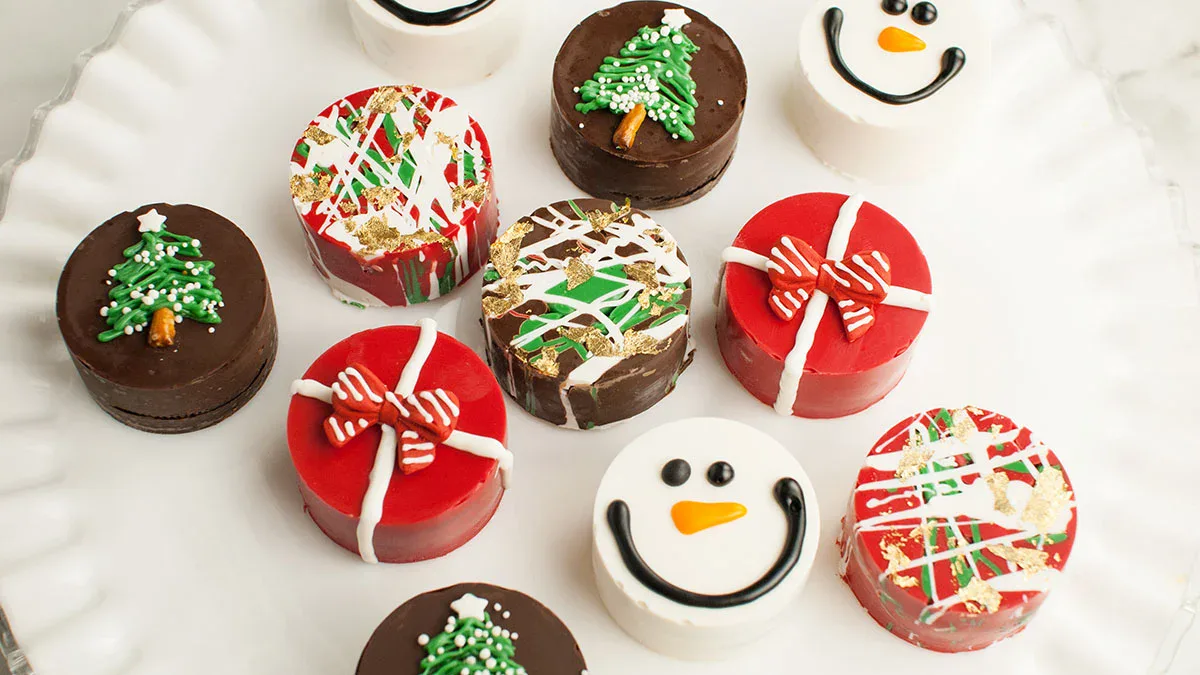 Assorted round holiday cookies decorated with Christmas trees, bows, snowflakes, colorful drizzles, and snowman faces on a white plate.