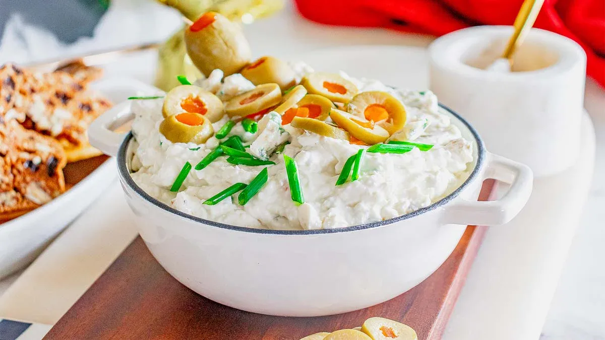 A white bowl filled with creamy dip, garnished with sliced green olives and chopped chives, sits on a wooden board.