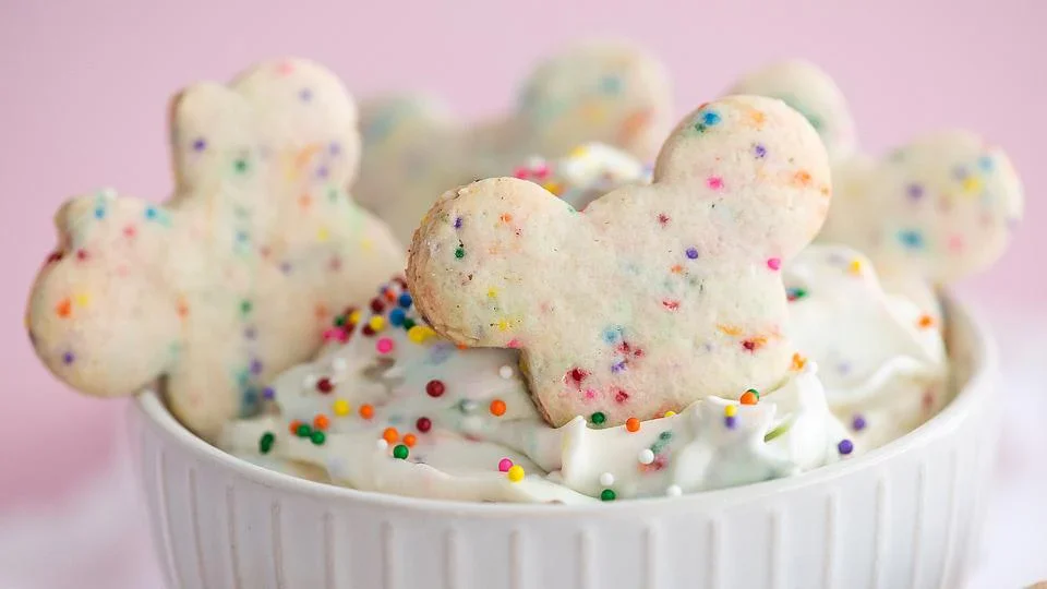 A white bowl filled with whipped cream, colorful sprinkles, and cookies shaped like Mickey Mouse heads with rainbow sprinkles.