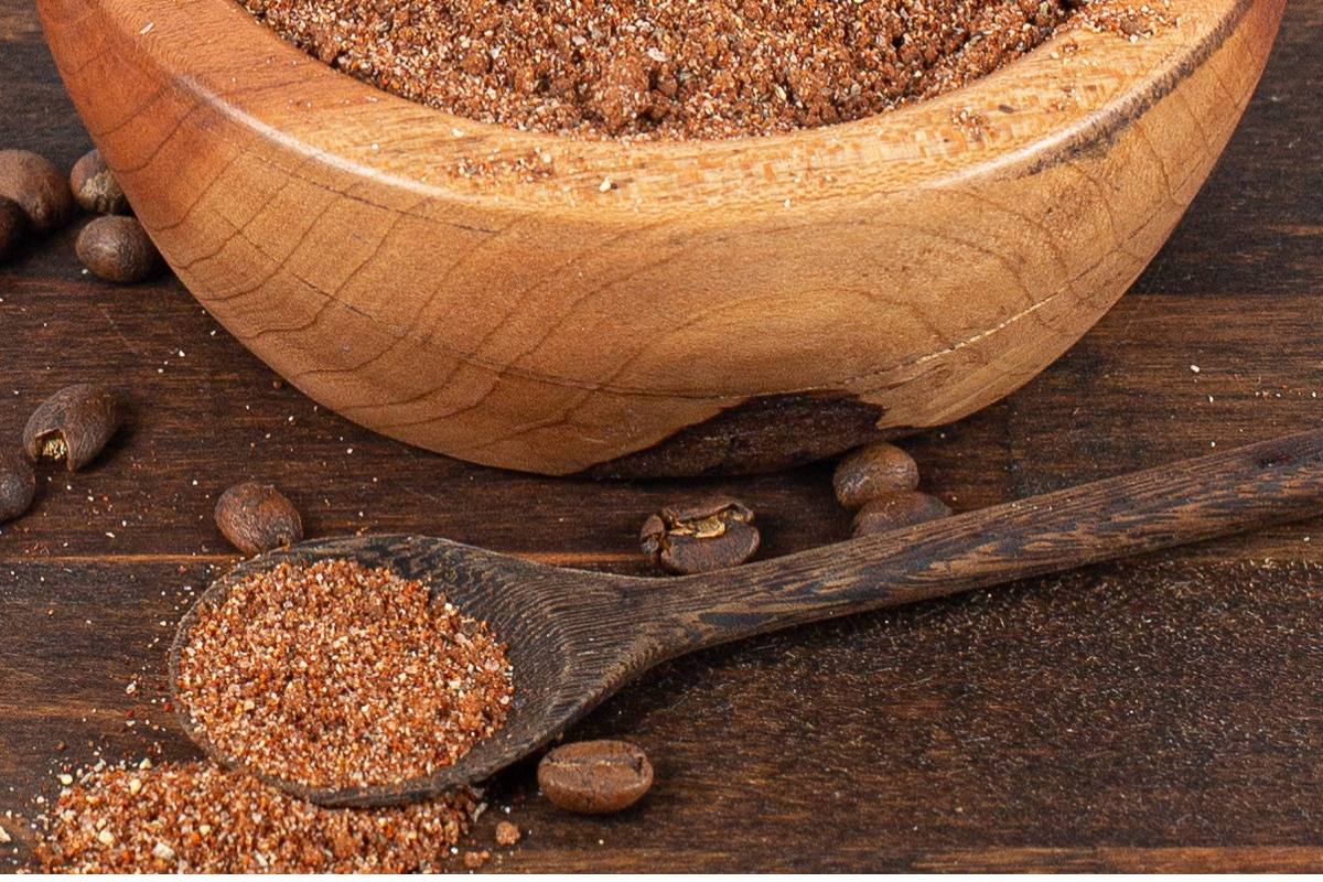 A wooden bowl filled with a ground brown spice mixture sits on a wooden surface, with a wooden spoon holding some of the mixture nearby and coffee beans scattered around.