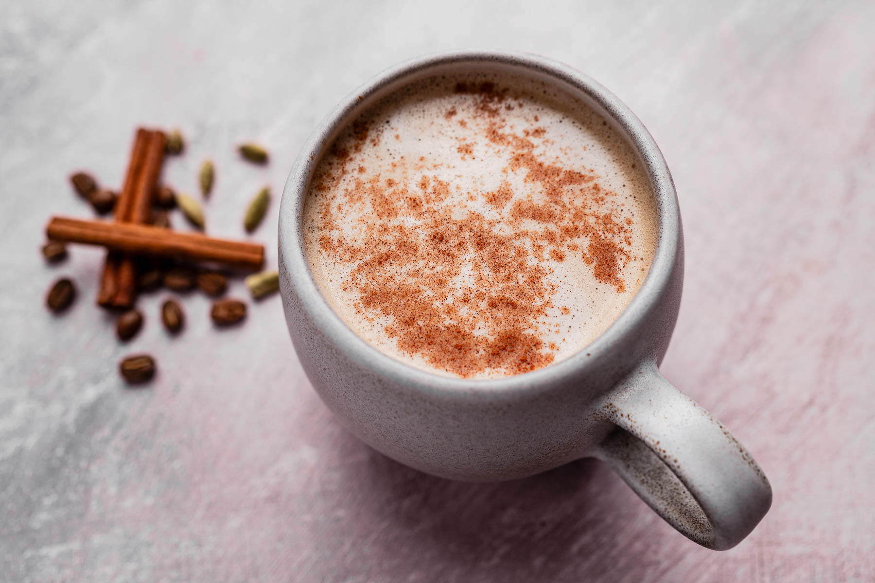 A ceramic mug filled with chai latte topped with cinnamon, placed on a light surface next to cinnamon sticks and cardamom pods.