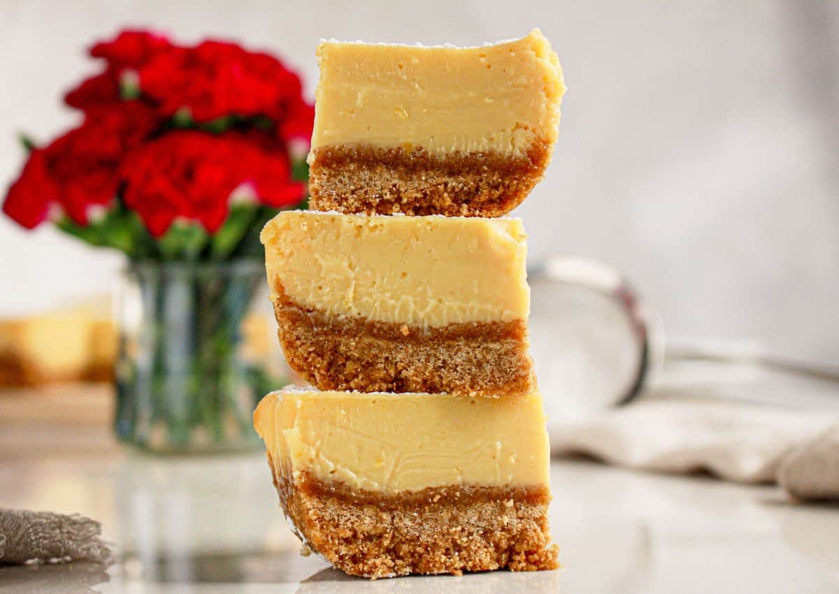 Three cheesecake bars with a crumbly base are stacked on top of each other on a countertop, with a jar of red flowers in the background.