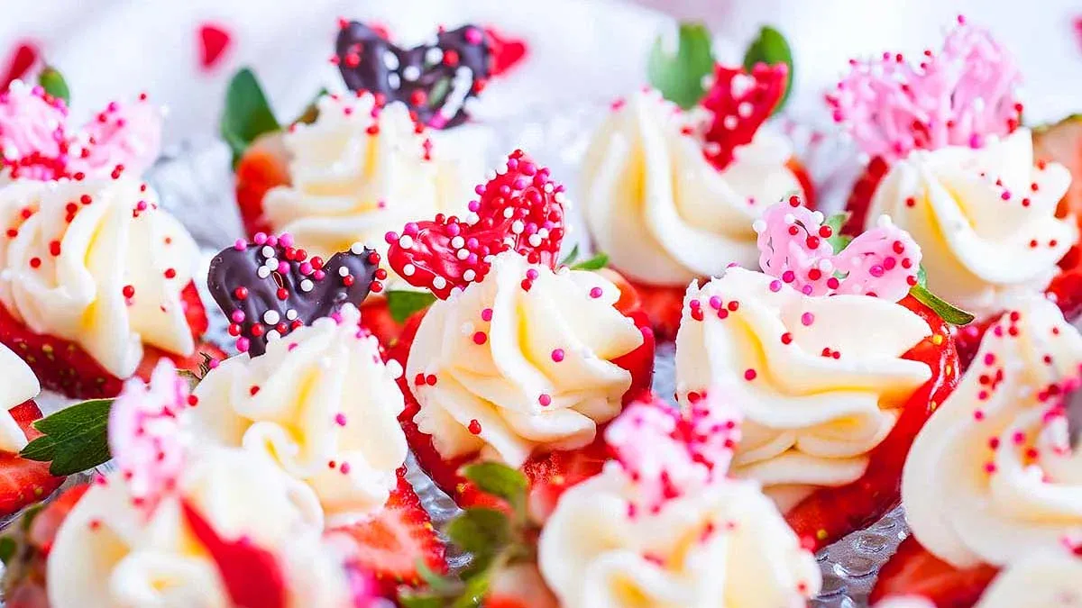 Rows of cupcakes topped with white frosting, pink and red heart-shaped decorations, and colorful sprinkles.