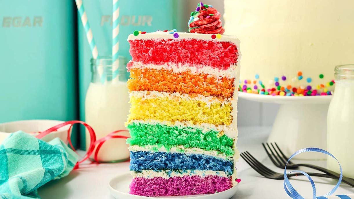 A slice of rainbow layer cake with white frosting and colorful sprinkles on a plate, next to milk bottles, forks, and pastel kitchen canisters.