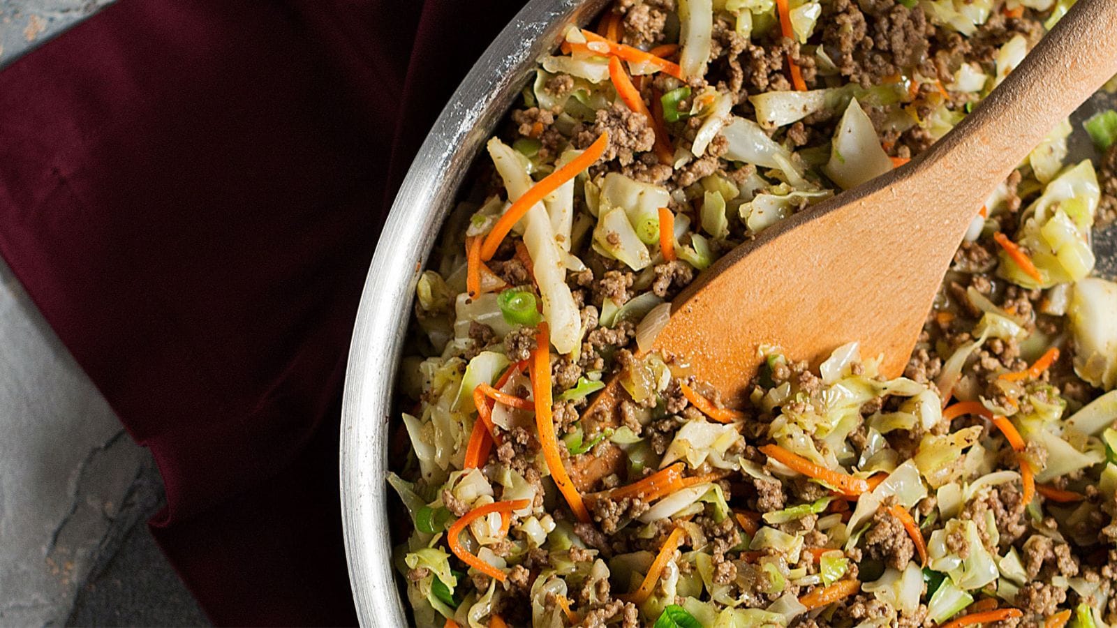 A skillet filled with cooked ground meat, shredded cabbage, carrots, and green onions, being stirred with a wooden spoon.