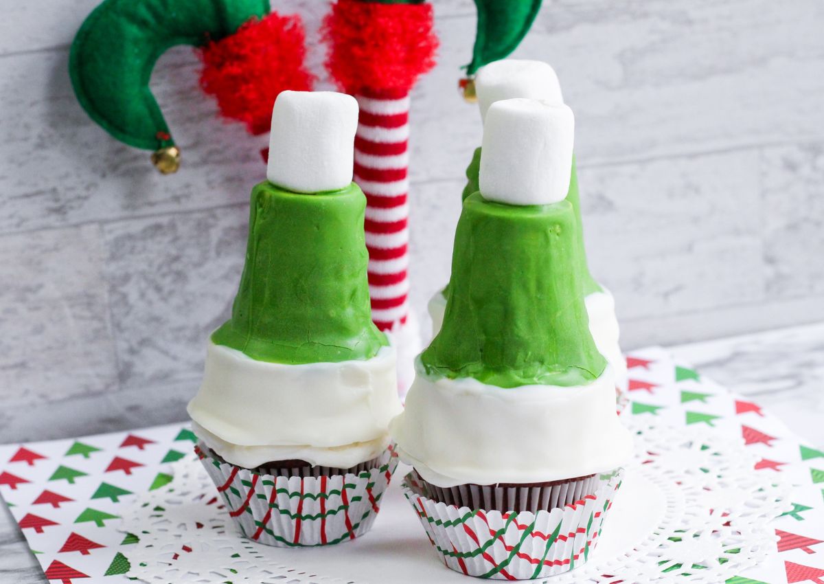 Three cupcakes are topped with green and white frosting, each holding an upside-down ice cream cone coated in frosting and topped with a marshmallow. Holiday-themed decorations appear in the background.