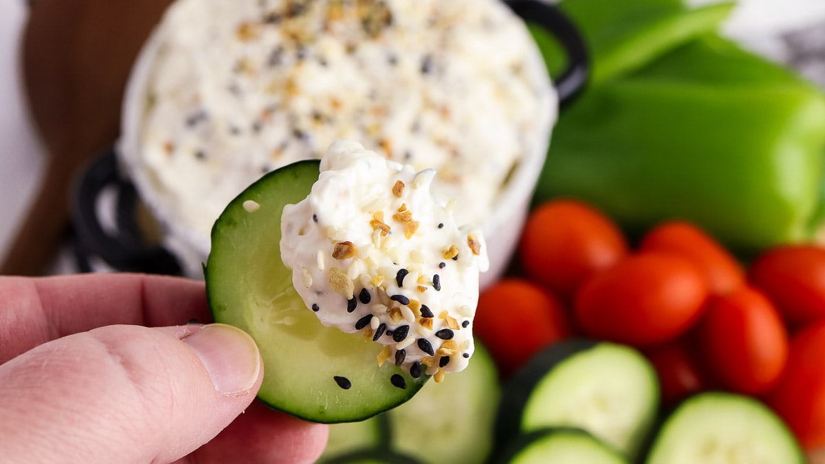A hand holds a cucumber slice topped with a creamy dip and seasoning, with sliced cucumbers, grape tomatoes, and green peppers in the background.