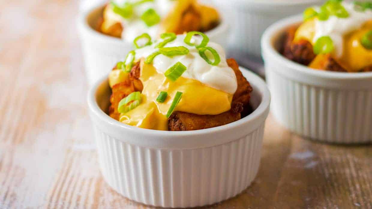 A small white ramekin filled with potato bites topped with melted cheese, sour cream, and sliced green onions.