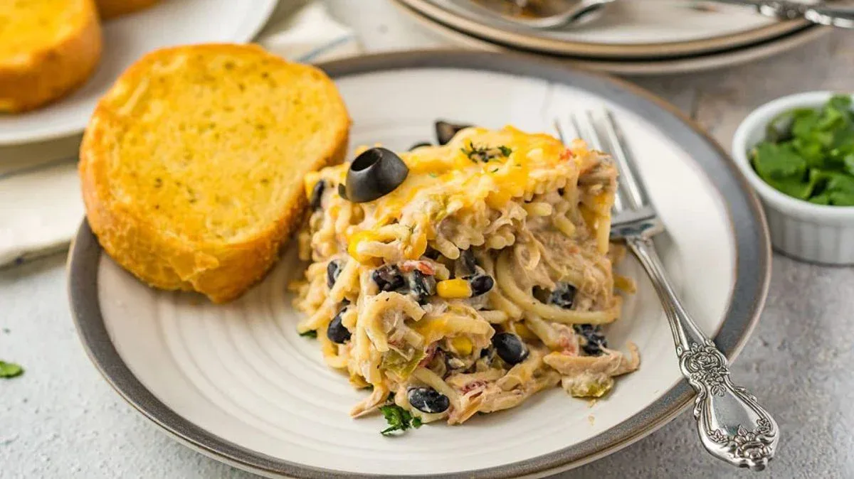A serving of cheesy baked pasta with black olives, corn, and shredded chicken on a plate, garnished with parsley, served with a slice of garlic bread.