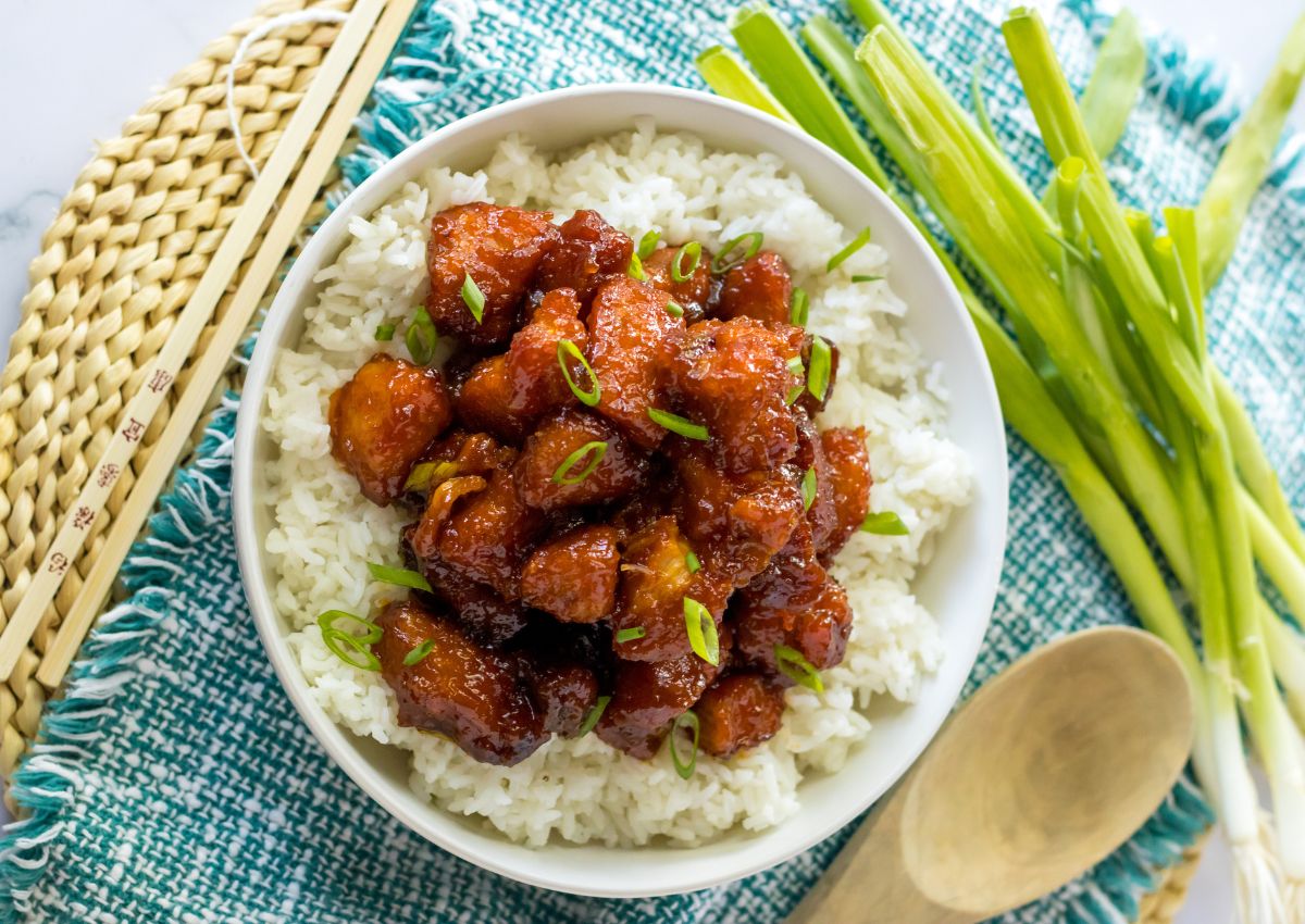 A bowl of white rice topped with glazed chicken pieces, garnished with sliced green onions, next to fresh green onions, chopsticks, and a wooden spoon.