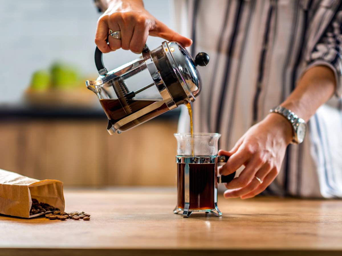 A person pours coffee from a French press into a glass mug on a wooden counter, with a bag of coffee beans nearby.