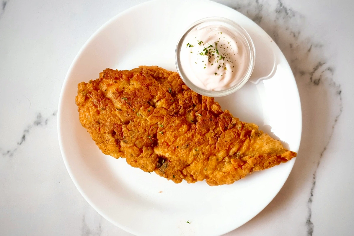 A piece of fried chicken breast on a white plate next to a small glass bowl of dipping sauce, garnished with herbs.