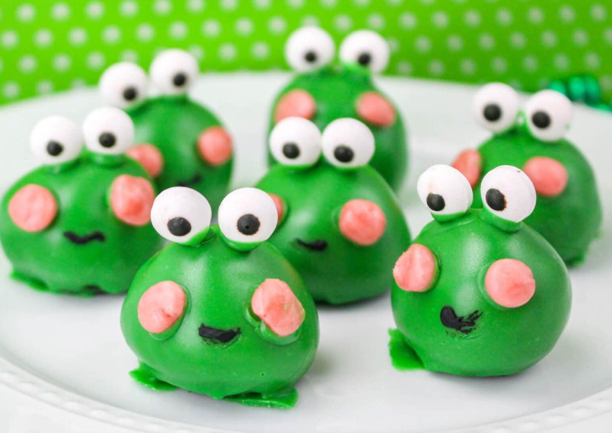 Seven green frog-shaped cake balls with large white eyes and pink cheeks are arranged on a white plate with a green polka dot background.
