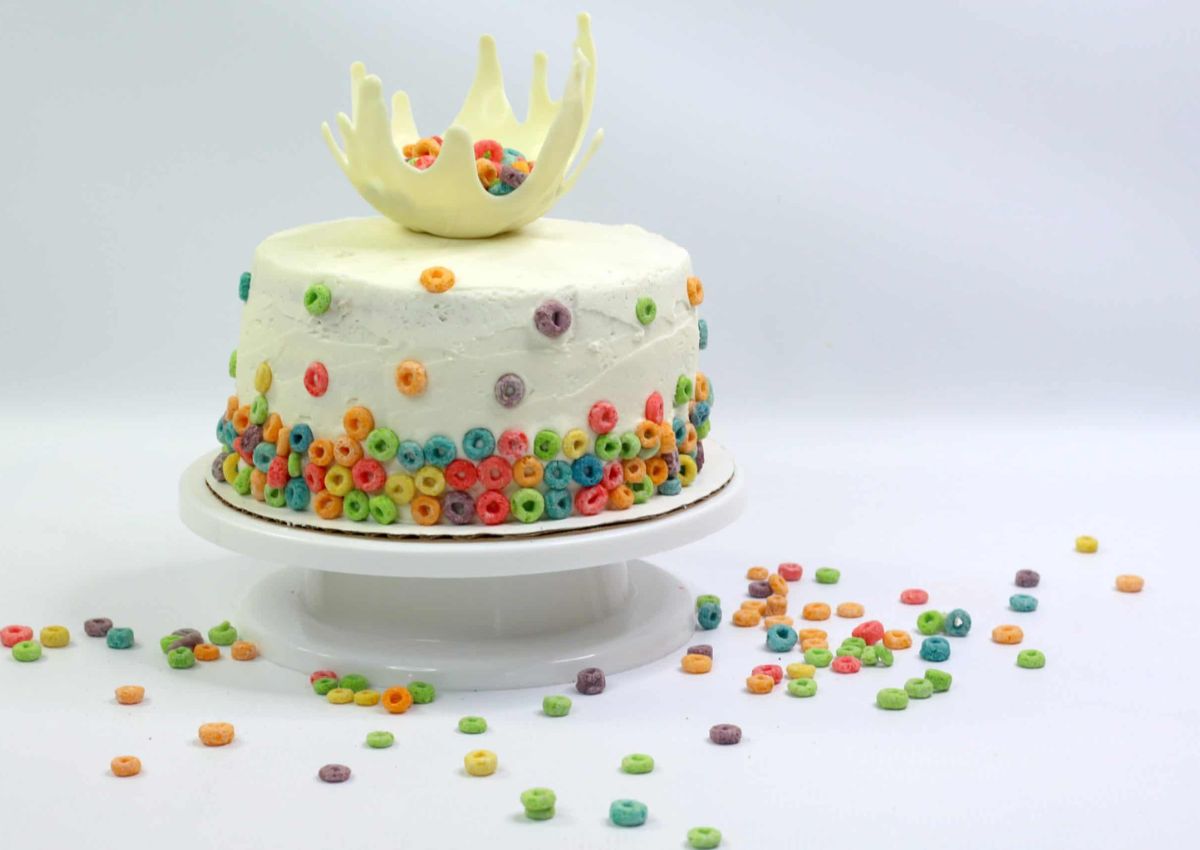 A round white frosted cake decorated with colorful cereal pieces and topped with a white chocolate crown, displayed on a white cake stand with cereal scattered around.