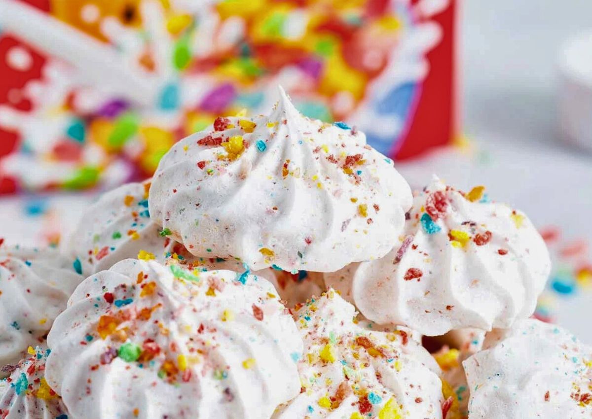 Close-up of white meringue cookies topped with colorful crushed cereal pieces, with a blurred cereal box in the background.