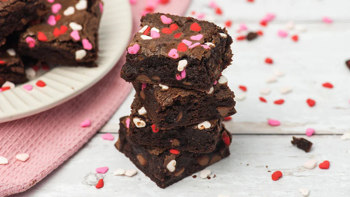 A stack of chocolate brownies with heart-shaped sprinkles on a white wooden surface, with more brownies and sprinkles on a plate in the background.