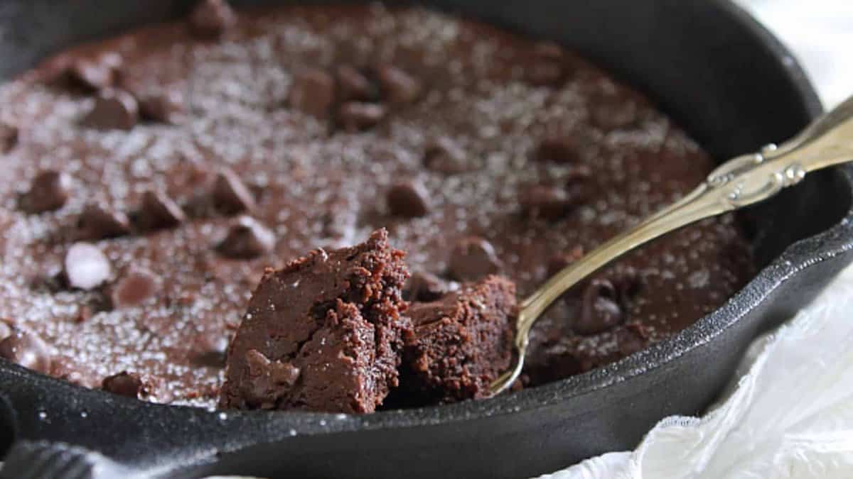A close-up of a skillet brownie topped with chocolate chips and powdered sugar, with a spoon lifting a portion from the pan.