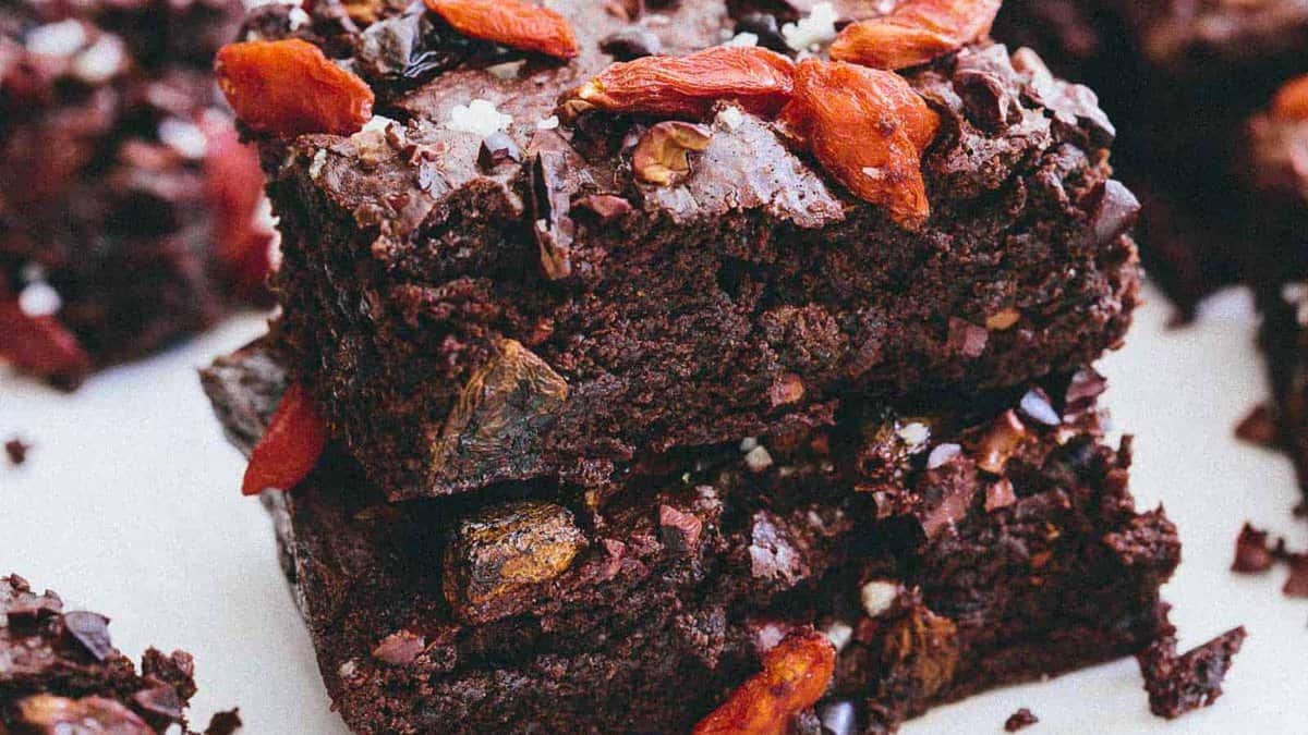Two stacked chocolate brownies topped with dried goji berries and coarse sea salt, with more brownies visible in the background.