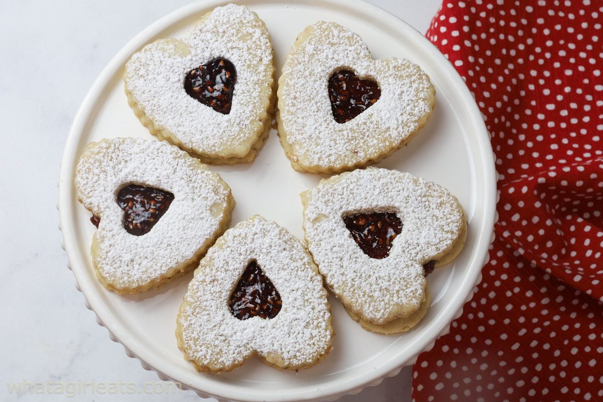 Six heart-shaped cookies with powdered sugar and jam centers are arranged in a circle on a white plate, next to a red and white polka dot cloth.